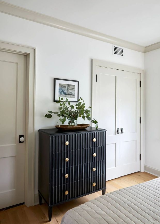 A bright, refined bedroom interior with white paneled walls and doors, traditional trim, pale timber flooring, and a black fluted chest of drawers topped with a leafy branch arrangement. The corner of a softly upholstered bed is visible in the foreground, confirming the room as a bedroom. A framed artwork and warm brass-toned drawer pulls add restrained decorative detail.