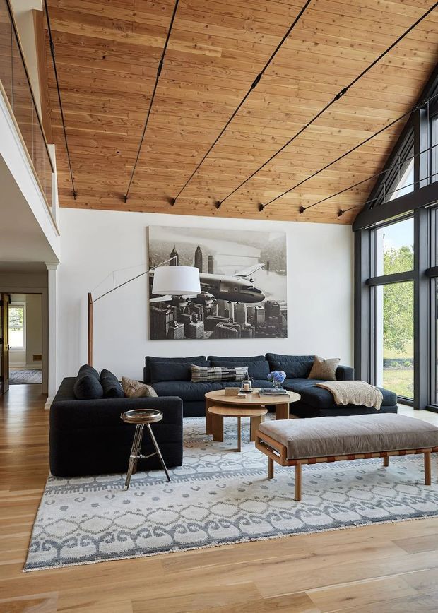 A spacious contemporary living room with a dramatic sloped ceiling clad in warm timber boards and exposed dark structural lines. The room features a large charcoal sectional sofa, black lounge chair, upholstered ottoman, round timber coffee tables, patterned area rug, and a monochrome city-and-aircraft artwork. Floor-to-ceiling black-framed glazing brings in abundant natural light and views of greenery, while an open passage connects to adjacent spaces.
