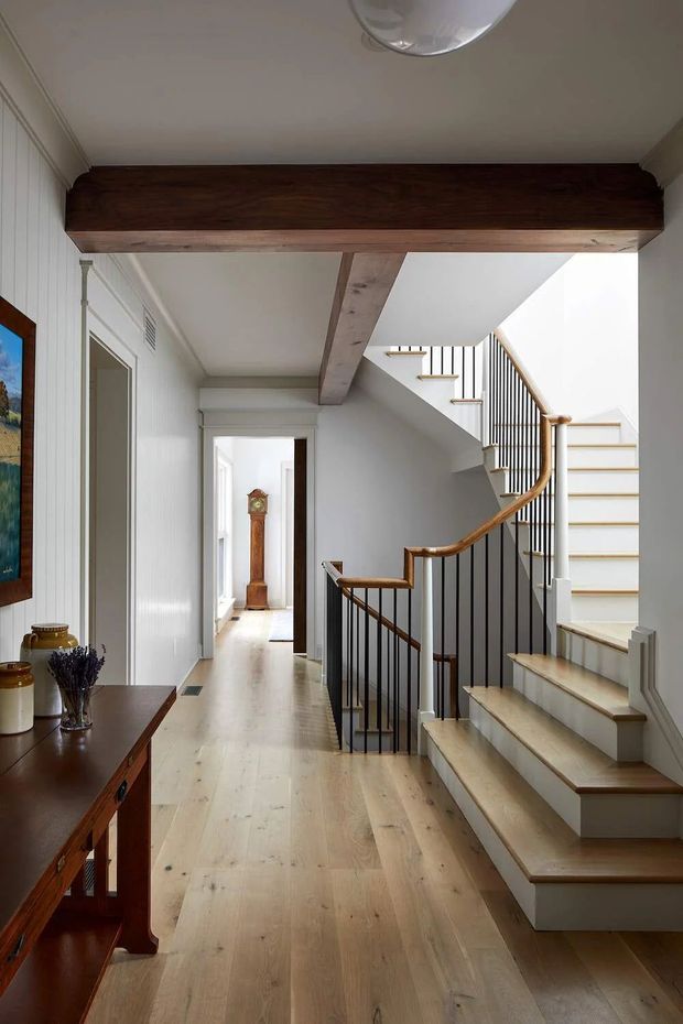 Bright traditional-style residential hallway featuring a timber staircase with white risers, dark wood handrails, and slender black metal balusters. Pale timber flooring runs through the hall, with white vertical wall panelling, decorative architraves, and a substantial exposed dark timber beam overhead. A console table and classic grandfather clock add traditional character.