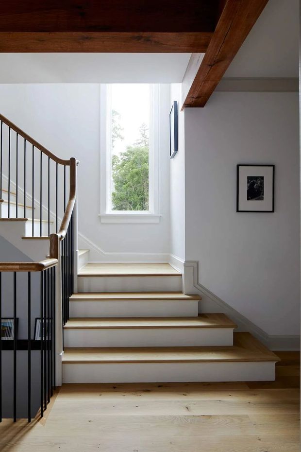 A bright residential stairway with pale timber flooring and stair treads, white-painted walls and risers, and a black metal balustrade with timber handrails. A tall window brings in natural light, while exposed dark timber ceiling beams and simple framed artwork add character.