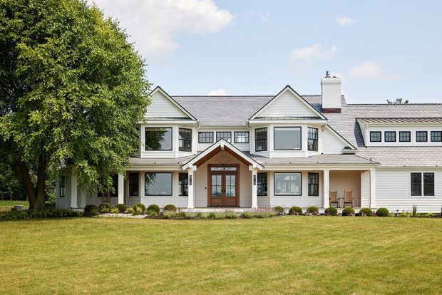 Front exterior view of a large, symmetrical two-storey suburban residence with white horizontal cladding, grey shingle-style pitched roofs, projecting gabled dormer-like sections, a central covered entry, and a long attached wing. The home is set behind a broad manicured lawn with mature landscaping.