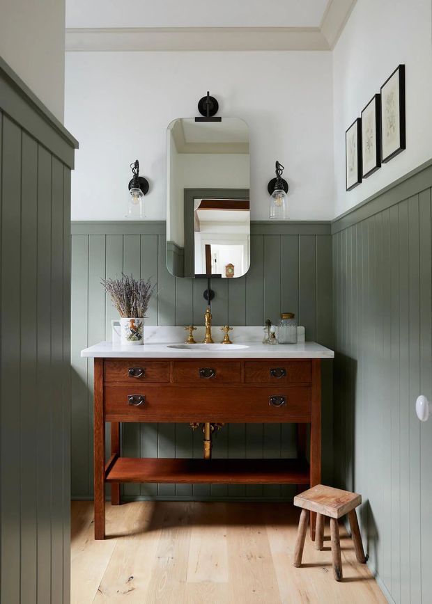 A compact, traditional-style bathroom with a vintage timber vanity, white countertop and basin, brass tapware, sage-grey vertical wall panelling, and pale timber flooring. A rounded rectangular mirror and twin wall sconces sit above the vanity, with framed artwork on the adjacent wall and a small wooden stool in the corner.