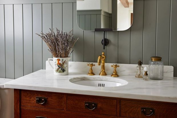 Close-up view of a traditional bathroom vanity with a white marble countertop, integrated oval basin, polished brass tapware, dark timber drawer cabinetry, and charcoal-grey vertical wall panelling. A rounded rectangular mirror and decorative accessories sit above the vanity.