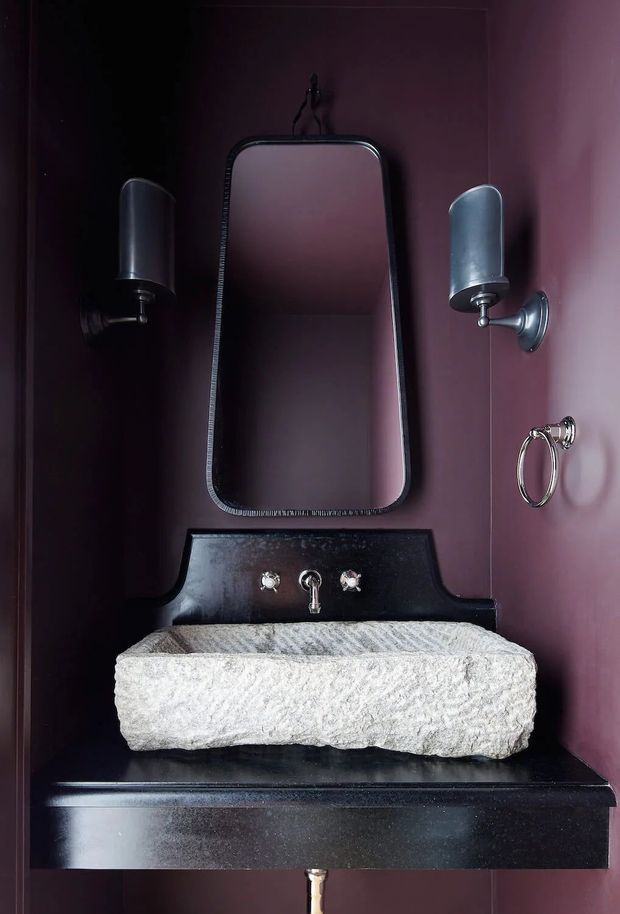 A compact, dramatic bathroom vanity vignette with a rough-hewn light stone basin set on a glossy black built-in counter. The deep burgundy painted walls are paired with dark metal or black-framed mirror, cylindrical wall sconces, polished metal tapware, and a ring towel holder.