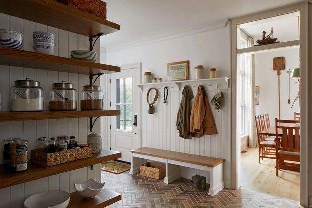 A bright, traditional-style home entryway with open timber shelving displaying crockery, glass jars, and pantry goods. A white vertical-panel wall has hooks holding coats and hats above a built-in timber bench with woven storage baskets. The space features a herringbone brick floor, white trim, and a glazed door leading to a dining area with timber furniture.
