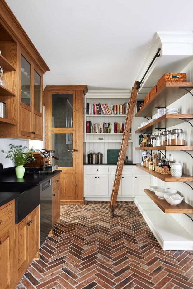 A compact, well-organised kitchen and pantry with warm timber cabinetry, white built-in shelving, open wooden storage shelves, black benchtops, and a striking reddish-brown herringbone brick floor. A timber ladder provides access to high-level shelving, while a central white cabinet wall displays books and kitchenware. The room has traditional detailing, including framed cabinetry, decorative cornices, and a glazed timber door.
