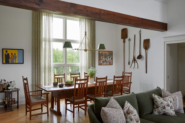 Bright open-plan dining and living area with a long timber dining table and matching wooden chairs positioned beneath a large exposed dark timber ceiling beam. Tall windows with sheer curtains provide abundant natural light. A green sofa with patterned cushions occupies the foreground, while eclectic wall art and decorative hanging wooden utensils add character.