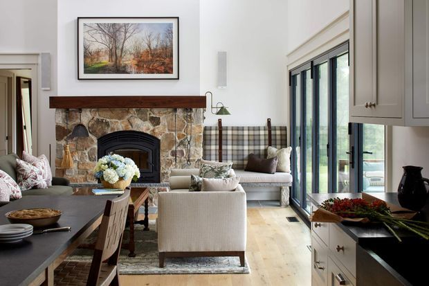 A refined open-plan living and dining space with a visible kitchen at right, centred on a rustic natural-stone fireplace with a dark timber mantel. The room combines light painted walls, pale timber flooring, dark kitchen surfaces, soft upholstered seating, patterned textiles, and tall black-framed glazed doors opening toward the outdoors. A framed landscape artwork and traditional built-in cabinetry add warmth and character.
