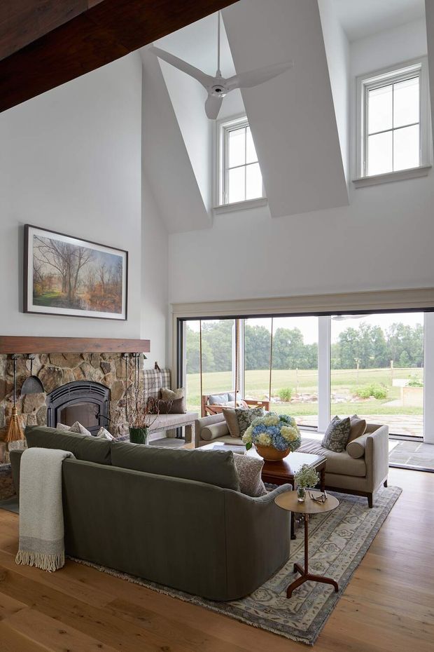 A bright, double-height living room with a vaulted ceiling, exposed dark timber beam, white walls, tall multi-pane windows, and expansive glazed doors opening to a rural landscape. The room features a substantial stone fireplace with timber mantel, a ceiling fan, timber flooring, a large patterned rug, and layered neutral upholstered seating arranged around a wooden coffee table.
