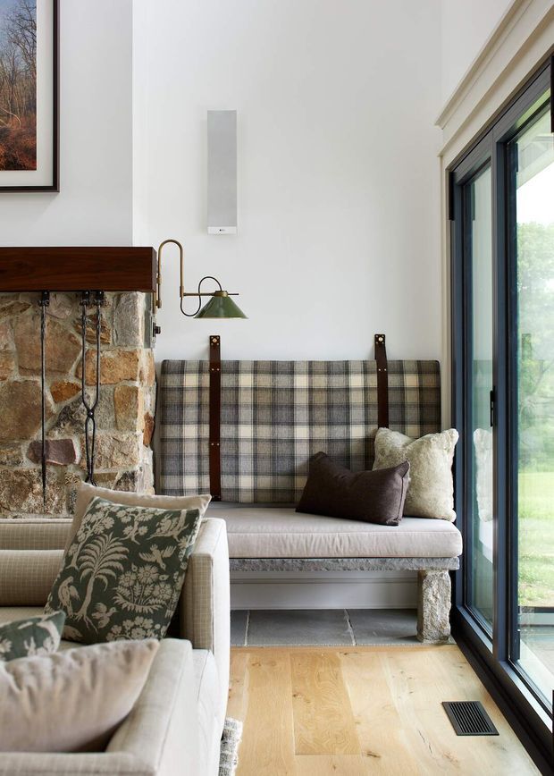 A cozy, light-filled sitting area with a built-in plaid upholstered bench secured by dark leather straps, layered neutral cushions, and a pale upholstered armchair. The room features a rough natural stone fireplace wall with timber mantel, white plaster walls, wide timber flooring, dark-framed full-height glazing, and a small wall-mounted reading light. The styling blends rustic natural materials with refined traditional furnishings.