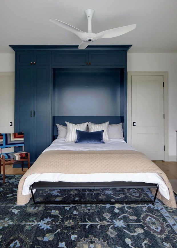 A well-appointed bedroom centered on a built-in dark navy bed wall with integrated wardrobes and overhead cabinetry. The bed has layered white and beige bedding with a navy accent cushion, set over a large patterned rug. A colorful upholstered chair sits to the side, while a white ceiling fan and paneled door complete the room.