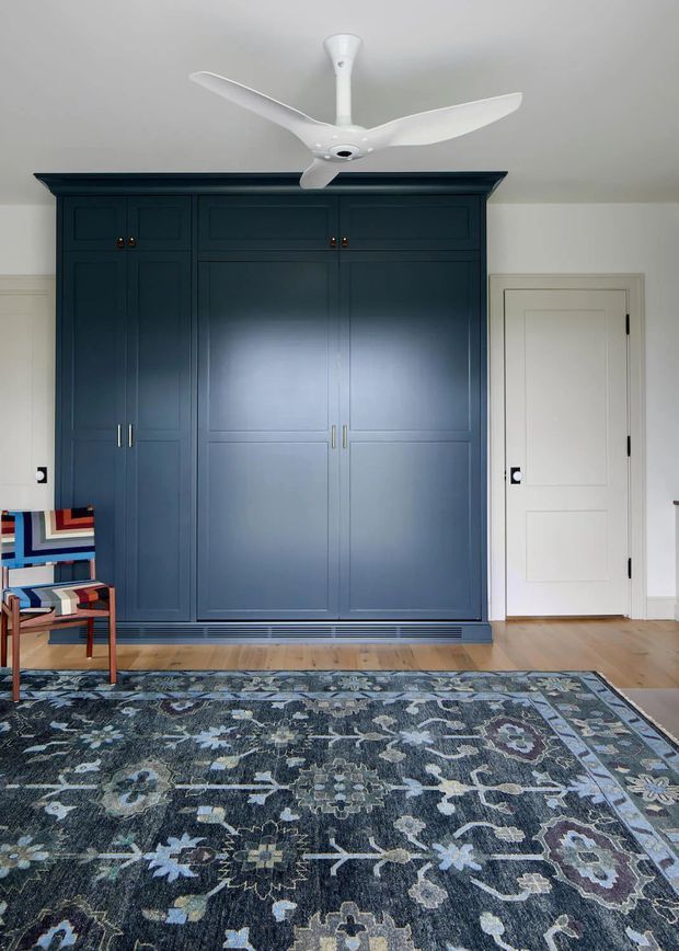 A refined bedroom interior featuring a full-height dark navy built-in wardrobe with paneled doors and brass handles, set against white walls. A white ceiling fan hangs above, while a white paneled door sits beside the wardrobe. Pale timber flooring and a large muted blue, grey, and cream patterned rug define the room, with a colourful upholstered occasional chair at the edge of the frame.