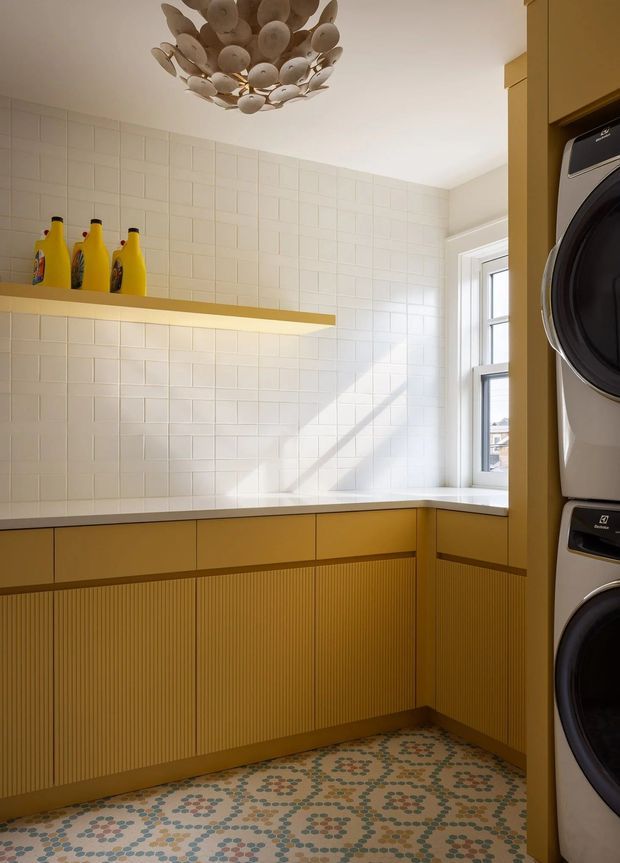 Bright, warmly styled laundry room with ochre mustard cabinetry, a white tiled wall, integrated countertop, open shelf with detergent bottles, stacked front-loading washer and dryer, and a decorative sculptural ceiling light. A patterned encaustic-style tile floor and large window add visual interest and natural light.