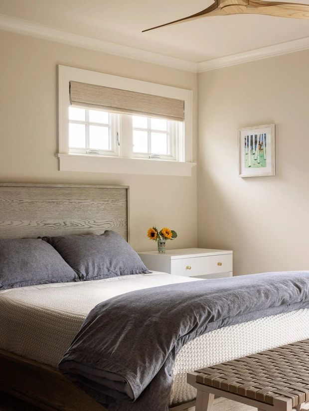 A serene, refined bedroom with a layered upholstered bed, light wood headboard, grey bedding, cream walls, white trim, and a small white bedside chest. A framed artwork, woven roller blind, decorative ceiling fan, and woven bench complete the warm neutral interior.