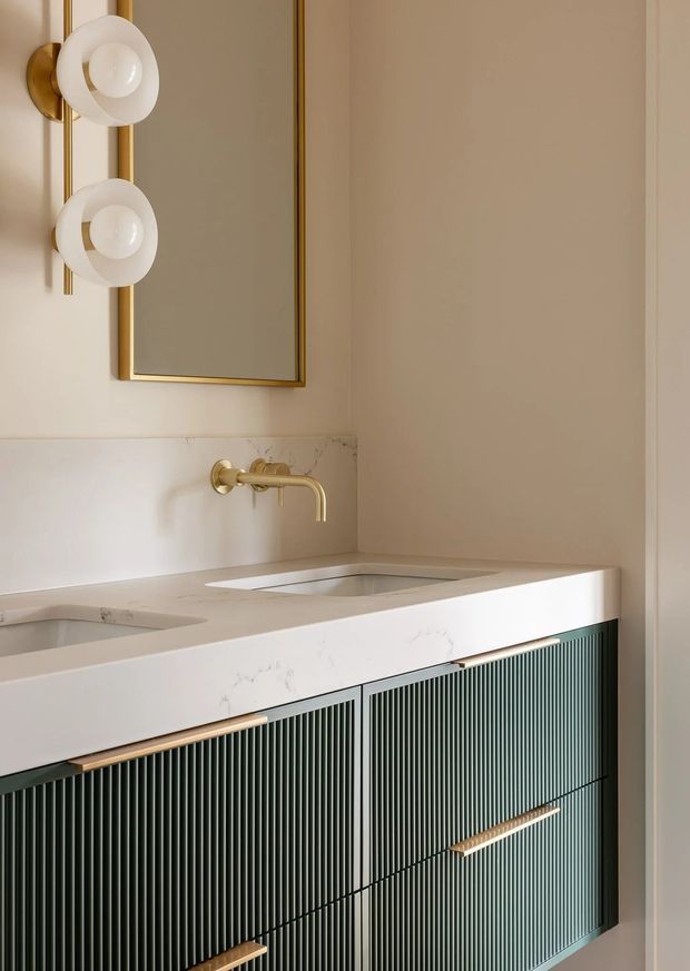 Close-up of a refined bathroom vanity featuring a double integrated basin in a pale veined stone benchtop, dark green vertically fluted cabinetry with slim brass handles, a wall-mounted brass tap, and a large brass-framed mirror. Sculptural white wall sconces add a decorative focal point against warm neutral walls.