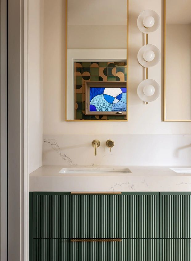 A refined bathroom vanity viewed straight on, featuring a fluted dark green built-in vanity with a white stone countertop and integrated basin. Brushed brass wall-mounted tapware and a vertical trio of rounded wall lights add warmth. A large brass-framed mirror reflects a colourful geometric tiled wall and a wall-mounted screen or artwork, creating a playful, art-deco-inspired focal point.