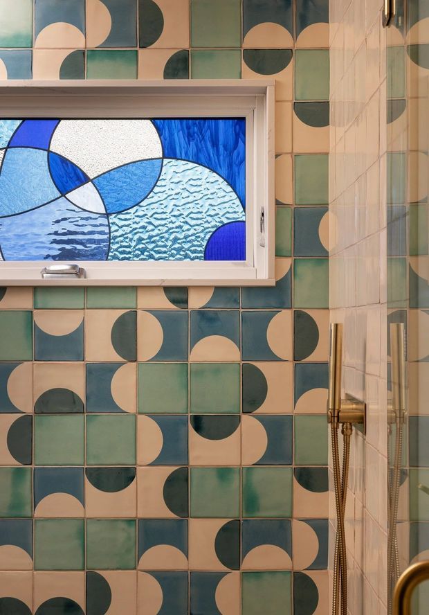 Close-up view of a richly tiled bathroom shower wall featuring a repeating geometric pattern of dusty teal, sage green, cream, and dark blue semicircles. A decorative stained-glass window with abstract blue and white water-like forms sits above the bath or shower area, while a glass shower screen and brass shower fittings are visible at right.
