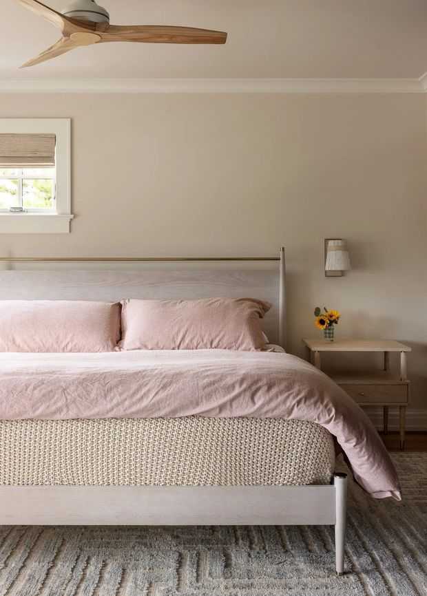 A softly styled bedroom with a pale timber bed featuring a woven-texture upholstered base, blush pink bedding and pillows, and a simple light-wood bedside table with a small lamp and flowers. The room has warm beige walls with decorative ceiling cornice, a small window with a woven blind, a timber ceiling fan, and a muted patterned grey rug.
