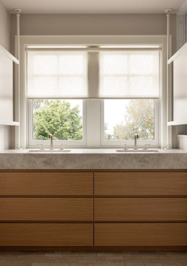 A refined bathroom vanity beneath a large double window, with warm wood drawers, a pale natural-stone benchtop, twin undermount basins and brushed metal tapware. Soft roller blinds filter daylight, while white shelving and trim frame the composition. A textured neutral wall and small-format floor tiles create a calm, understated interior.