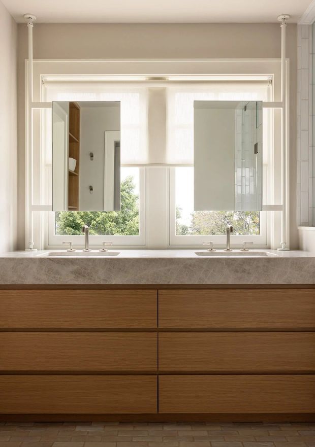 A refined bathroom vanity with a wide double-sink stone countertop, warm horizontal-grain timber cabinetry, twin wall-mounted mirrors, and brushed metal faucets. Large divided windows provide abundant natural light, while slim white structural trim and pale neutral walls create a calm, balanced composition. A glass shower enclosure is partially visible at the right edge.