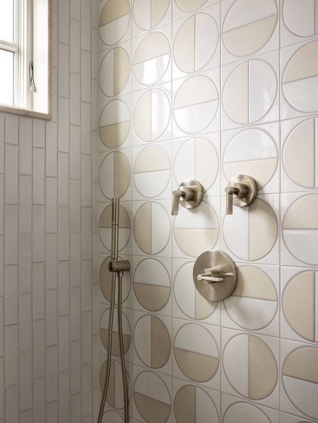 Close-up view of a refined shower area with a distinctive geometric tiled feature wall composed of overlapping circles and semicircles in soft white and beige tones. Brushed metallic shower controls, a handheld shower rail, and a small window are visible. The space is bright, clean, and subtly decorative.
