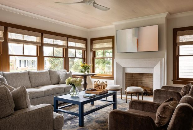 A warm, traditional living room with large timber-framed windows, woven blinds, a grey sectional sofa and armchairs, a dark coffee table, patterned area rug, and a white textured fireplace surround with exposed brick firebox. A wall-mounted television sits above the fireplace, while a ceiling fan and timber ceiling add to the relaxed residential setting.