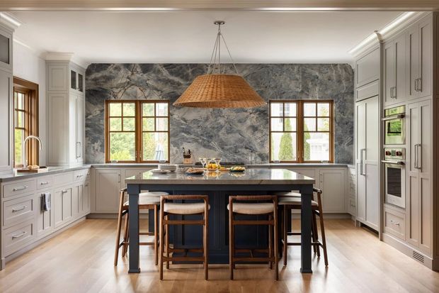 A refined, spacious kitchen with a central island and four bar stools, framed by pale grey built-in cabinetry and integrated appliances. The island has a dark painted base and a light stone benchtop, while the rear wall features dramatic grey veined stone slab cladding. Timber-framed windows bring in natural light, and a large woven pendant adds warmth above the island. The room has timber flooring and classic detailed cabinetry with crown moulding.