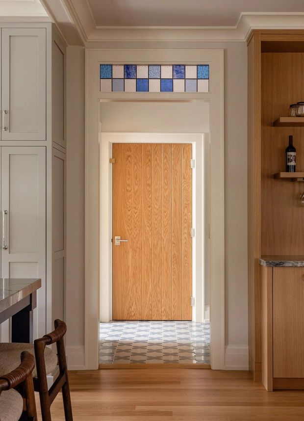 A refined interior view centred on a timber door framed by painted panelled joinery and a decorative transom of small blue, cream and grey glass tiles. The surrounding space includes built-in cabinetry, a timber and stone-look bar or display unit, partial dining chairs, and a patterned tiled threshold transitioning to timber flooring. The room has warm neutral finishes and traditional detailing.