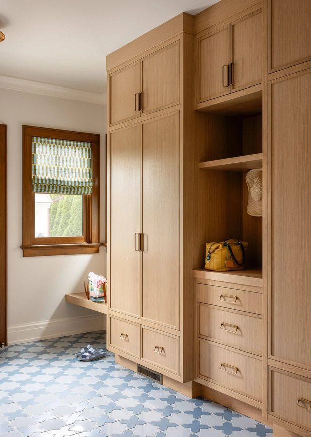 A bright, traditional-style mudroom or entry storage area with extensive floor-to-ceiling warm wood cabinetry, framed doors, drawers, open shelving, and dark metal handles. A patterned blue-and-white tile floor, wood-trimmed window with woven Roman shade, built-in bench, and small personal items complete the space.