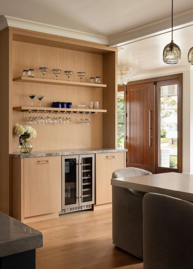 A warm, refined open-plan entertaining area featuring a built-in timber bar and drinks cabinet with open glassware shelving, under-counter wine refrigeration, and a dark stone benchtop. The space connects to a dining area with upholstered seating and includes a timber-framed glazed entry door, pendant lighting, and traditional ceiling mouldings.