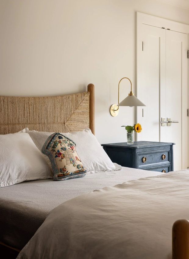 A warm, softly lit bedroom with a woven natural-fibre bedhead, white bedding, a dark blue bedside chest, brass wall sconce, white paneled wardrobe doors, and a decorative patterned cushion. The room has a relaxed, refined coastal feel with warm neutral finishes and natural textures.
