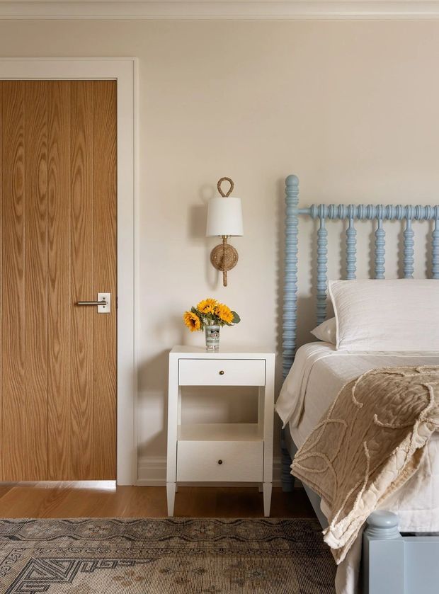 A warm, refined bedroom corner featuring a white bedside table, blue turned-post bed frame, cream bedding, a timber door, wall-mounted lamp, patterned rug, and soft neutral finishes.