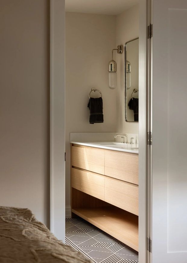 View from a bedroom through an open door into a compact, refined bathroom. The bathroom features a floating light-wood vanity with integrated drawers, a white benchtop and basin, brushed metal tapware, a wall-mounted mirror, glass wall sconces, and a patterned geometric tile floor. A towel ring with dark towels is mounted beside the vanity. Part of the bedroom and bed is visible in the foreground, clearly showing the bathroom connection.