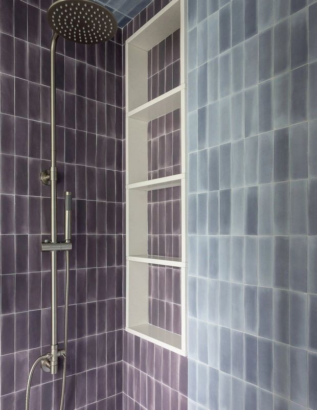 Close-up view of a tiled shower enclosure with dark purple ceramic wall tiles, pale blue tiled side wall, a white recessed vertical niche with multiple shelves, and a dark metal rainfall shower system with handheld attachment.