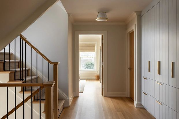 A bright, refined residential hallway viewed from beside a staircase. The space features warm timber flooring, a painted stair with timber handrails and black metal balusters, white paneled built-in cabinetry with brass-toned handles, painted walls and trim, and a doorway leading toward a naturally lit room.