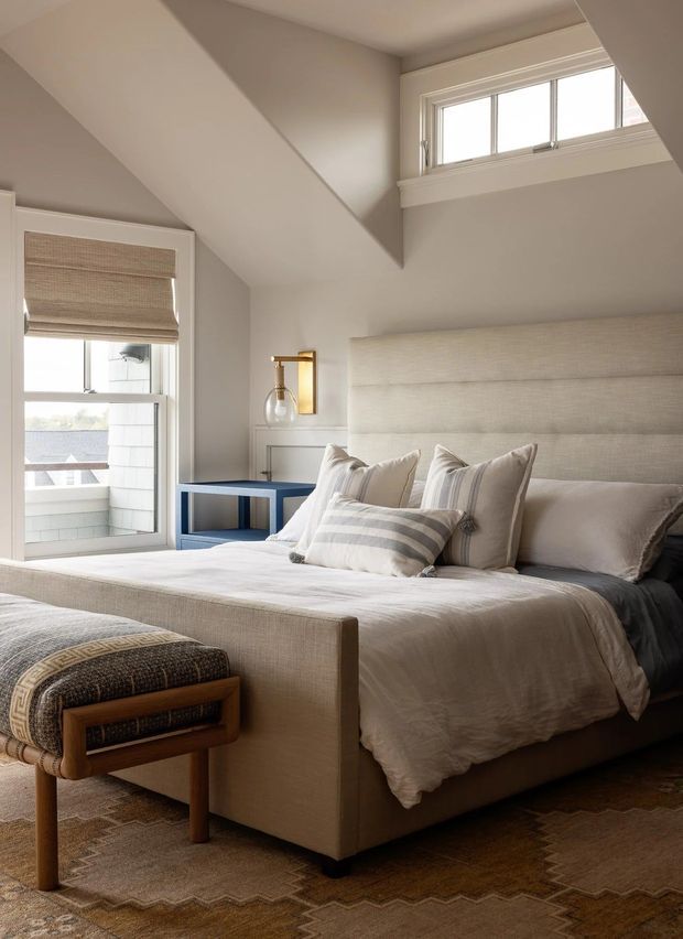 A refined, light-filled bedroom with a large upholstered bed, layered neutral bedding, a padded wall-to-wall headboard, bedside table, wall sconce, woven window shades, and a patterned area rug. High-level windows and an angled ceiling create an airy, tailored atmosphere with warm beige, cream, and soft grey tones.