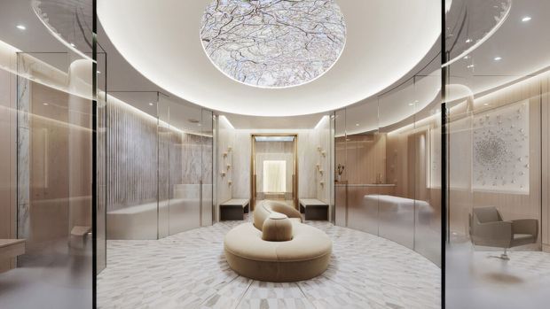 A luxurious, symmetrical interior lobby or reception area with curved mirrored and glass walls, pale stone-patterned flooring, softly illuminated coved ceiling, and a large circular sculptural ceiling light. A rounded upholstered central seating island anchors the space, with additional lounge seating and refined light-toned wall finishes surrounding it.