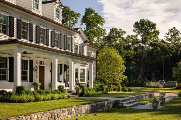 Exterior view of a large, symmetrical white clapboard-style residence with black shutters, multi-pane windows, dormer windows, and a covered entrance portico supported by classical columns. The landscaped grounds include manicured lawns, clipped shrubs, a low stone retaining wall, steps, and a formal garden path surrounded by mature trees.