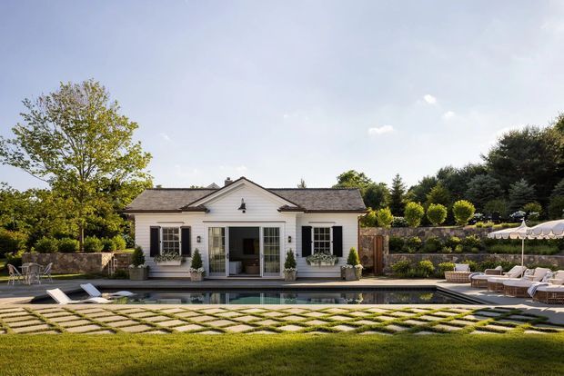 A single-storey white pool house or pavilion with a symmetrical gabled facade, dark shutters, timber or dark-framed glazed doors, and stone masonry side walls. It fronts a long swimming pool with surrounding patterned paving, manicured lawn, clipped shrubs, mature trees, and outdoor lounge furniture. The setting is refined and resort-like, with traditional architectural detailing and a calm garden landscape.