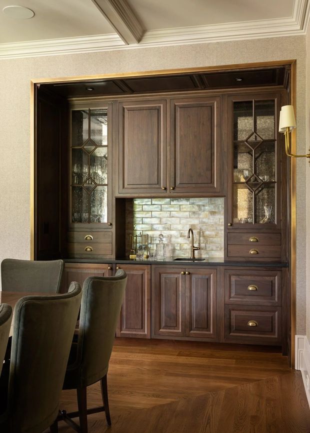 A refined traditional dining area featuring a dark stained timber built-in bar or kitchenette, with framed cabinetry, glass-front display cupboards, brass hardware, a small sink, and a pale textured brick-style splashback. Dark upholstered dining chairs sit in the foreground. The room has warm timber flooring, patterned wallcovering, and ornate white coffered ceiling trim.