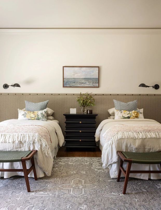 A refined twin bedroom with two neatly made beds positioned symmetrically around a black chest of drawers. The room features a padded upholstered wall-to-wall headboard, cream bedding with fringed throws, patterned accent cushions, brass-and-black wall sconces, a framed coastal artwork, dark timber flooring, and a large muted patterned rug. Two upholstered timber-framed stools sit at the foot of the beds.