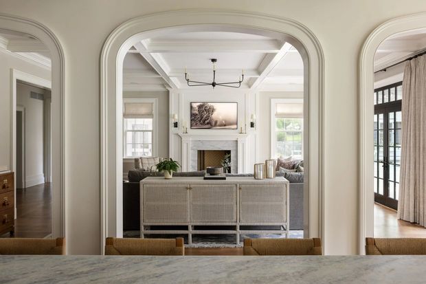 Elegant view through a series of arched openings into a refined living room, with a white coffered ceiling, classical fireplace surround, pale walls, dark timber flooring, large windows, and a woven-front console. The foreground appears to be a stone-topped dining or kitchen counter, while a glazed sunroom or enclosed terrace is visible to the right.