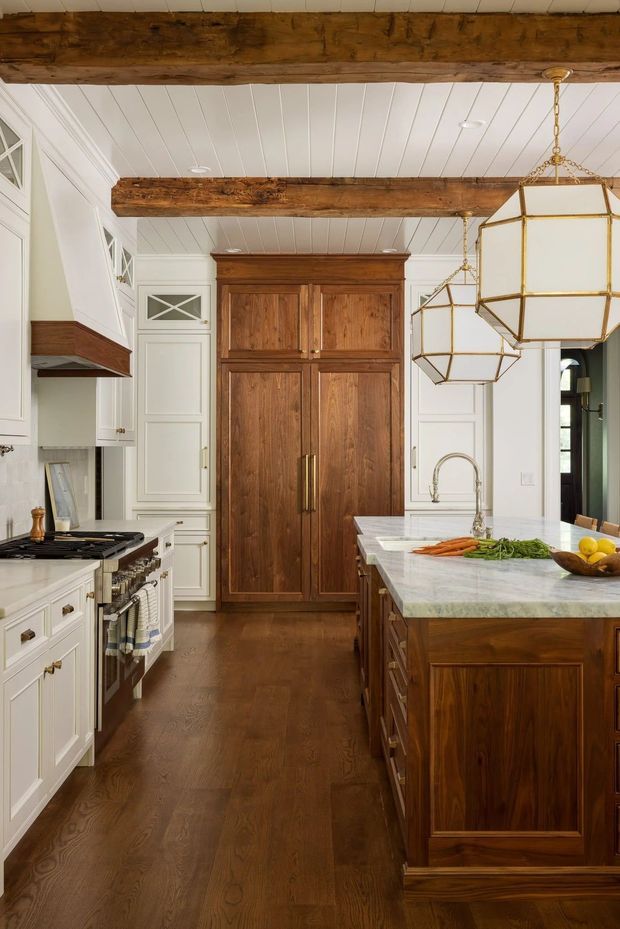A refined traditional kitchen with white paneled cabinetry, rich walnut-toned island and tall pantry cabinetry, dark timber flooring, exposed rustic wood ceiling beams, and a white beadboard ceiling. The kitchen features a pale stone island benchtop, brass hardware, a large range, farmhouse-style sink tapware, and striking geometric brass-and-glass pendant lights.