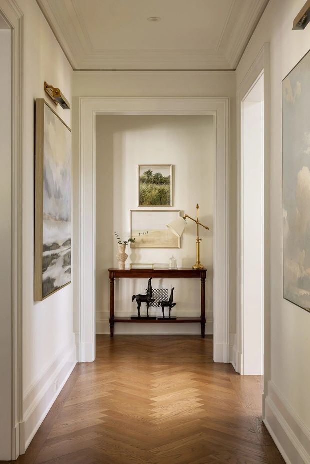 A refined residential hallway with warm herringbone timber flooring, cream-painted walls, detailed white wall and ceiling mouldings, and wide decorative door trim. The corridor opens to a small display alcove containing a dark wood console table, brass floor lamp, framed artwork, sculptural decor, and a pair of black chairs. Large framed artwork hangs along both sides of the passage, creating a gallery-like, classically detailed interior.