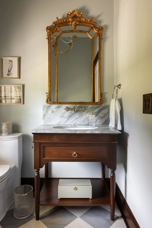 A refined traditional bathroom vanity vignette featuring a dark timber console-style vanity with turned legs, a white marble benchtop and integrated marble backsplash, brass plumbing fixtures, and an ornate gilt-framed mirror. Pale grey walls, patterned grey-and-white floor tiles, framed artwork, towel hooks, and a toilet complete the compact bathroom.