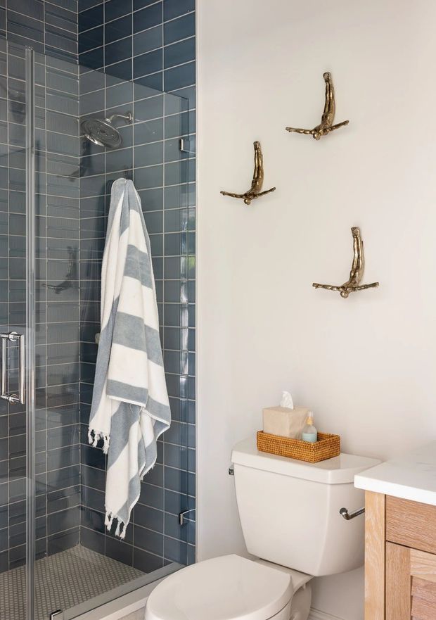 A compact contemporary bathroom featuring a glass-enclosed walk-in shower lined with blue-grey rectangular tiles, a small patterned tile floor, a white toilet, and a light timber vanity. Decorative brass wall hooks and a striped towel add warmth and detail.