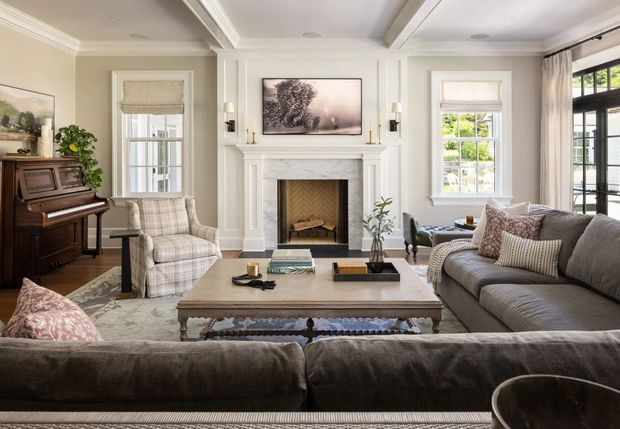 A refined traditional living room with a large upholstered sectional sofa arranged around a substantial central coffee table and a white classical fireplace mantel. The room features symmetrical wall panelling, decorative ceiling beams and crown moulding, tall sash-style windows with Roman shades, a piano, patterned armchairs, and neutral layered textiles. French doors open toward a bright exterior.