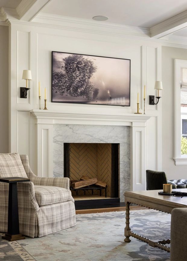Elegant traditional living room centred on a white painted fireplace with a marble surround and herringbone brick firebox. The room features detailed wall panelling, layered crown moulding, black wall sconces, a large framed artwork above the mantel, a patterned upholstered armchair, and a turned-leg coffee table over a muted area rug.