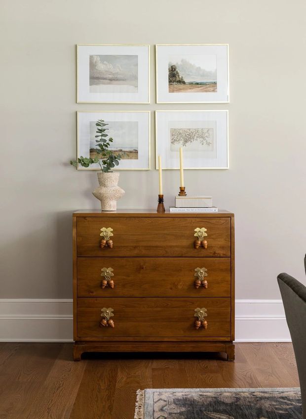 A warm, traditionally styled interior vignette featuring a dark wood three-drawer chest with ornate brass-and-wood pulls. The chest sits against a soft grey wall with white trim, topped by a ceramic vase with greenery, two taper candles, and stacked books. Four delicate framed landscape artworks hang above it, with timber flooring and part of an upholstered chair and rug visible at the edges.