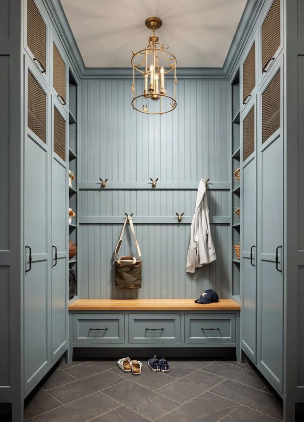 A compact, highly fitted mudroom or walk-in storage space with symmetrical blue-grey cabinetry, woven rattan-style door panels, open shelving, a central timber bench with drawers, coat hooks, and vertical wall panelling. A brass cage-style pendant light hangs from the moulded ceiling, while dark large-format floor tiles create contrast. Shoes, a bag, coat, and cap reinforce the entry and storage function.